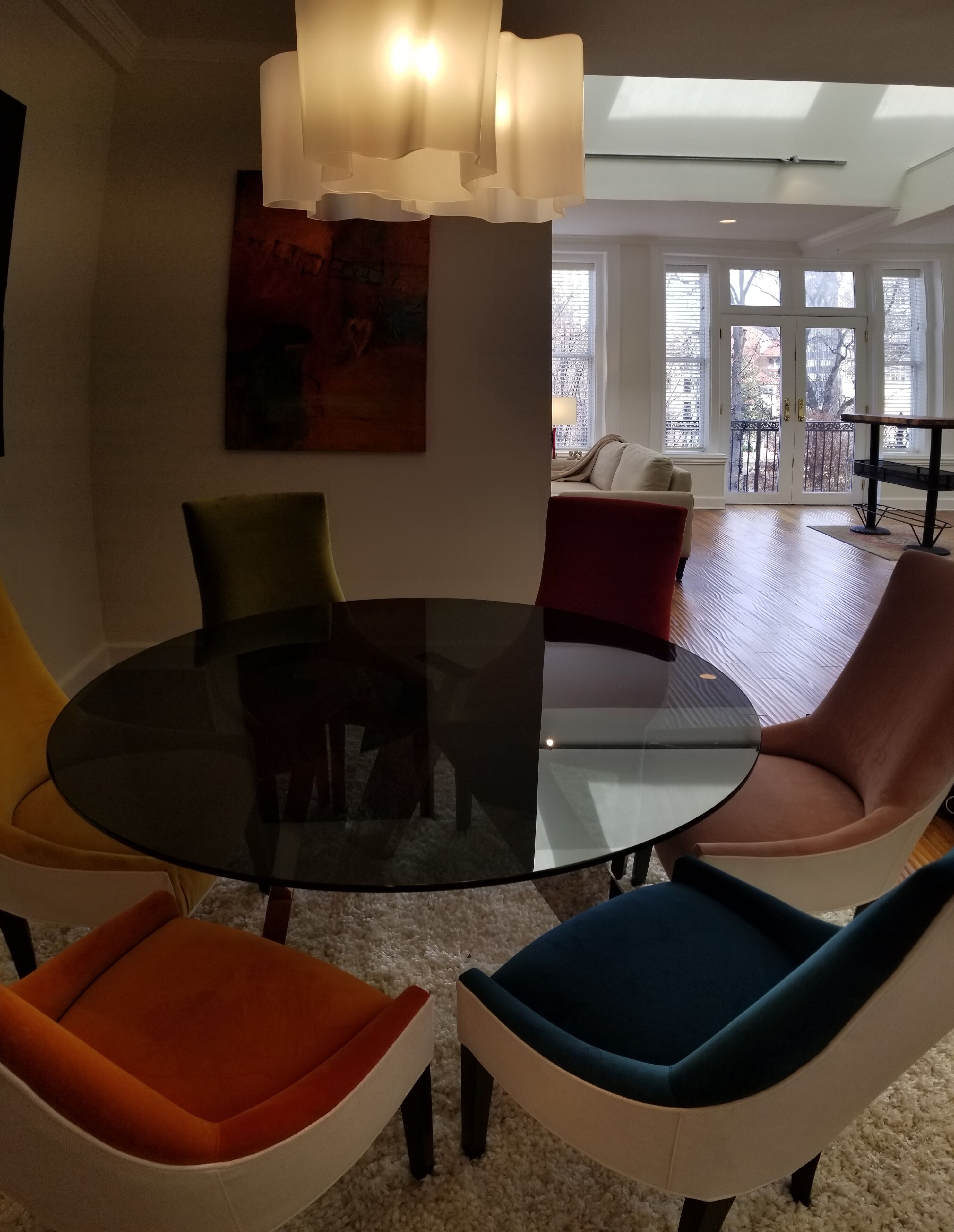 Una sala de estar moderna con una mesa de centro de vidrio rodeada de sillas coloridas, un cuadro grande en la pared y vistas de la ciudad a través de amplias ventanas.