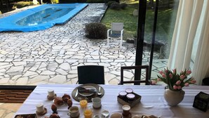 Una mesa con desayuno que incluye cruasanes, pasteles, fruta y bebidas, con vistas a una piscina y un jardín.