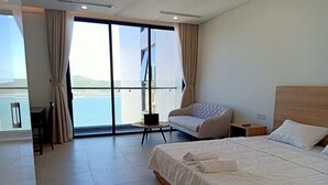 A modern hotel room with a large bed, a sofa, a small table, and a view of the sea through large windows.