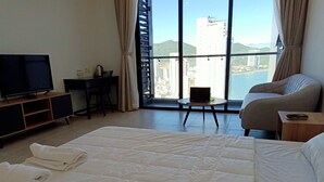 A hotel room with a large bed, a desk, a chair, a TV, and a view of a cityscape through large windows.