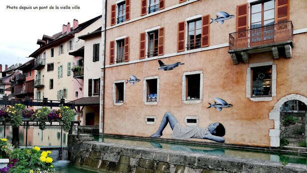 A canal with a mural of a person lying on a wall, buildings with balconies, and a bridge with flowers. - Large studio in the center of Annecy (Annecy)