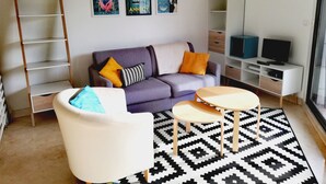 A modern living room with a sofa, a white armchair, a coffee table, a TV stand, and a bookshelf. There are three posters on the wall. - Large studio in the center of Annecy (Annecy)