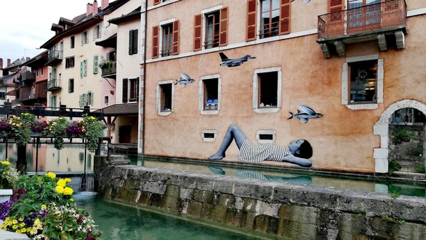 A canal with a mural of a person lying on a wall, buildings with balconies, and a bridge with flowers.