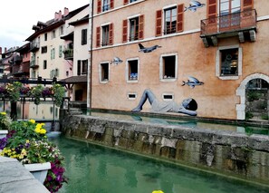 A canal with a mural of a person lying on a wall, buildings with balconies, and a bridge with flowers.