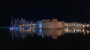 Un muelle histórico con un edificio fortificado, agua tranquila que refleja las construcciones, y un puerto deportivo con veleros.