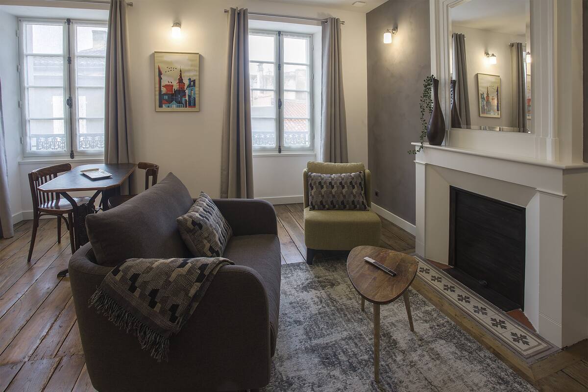 A living room with a sofa, armchair, and small table. There is a fireplace and a window with curtains.