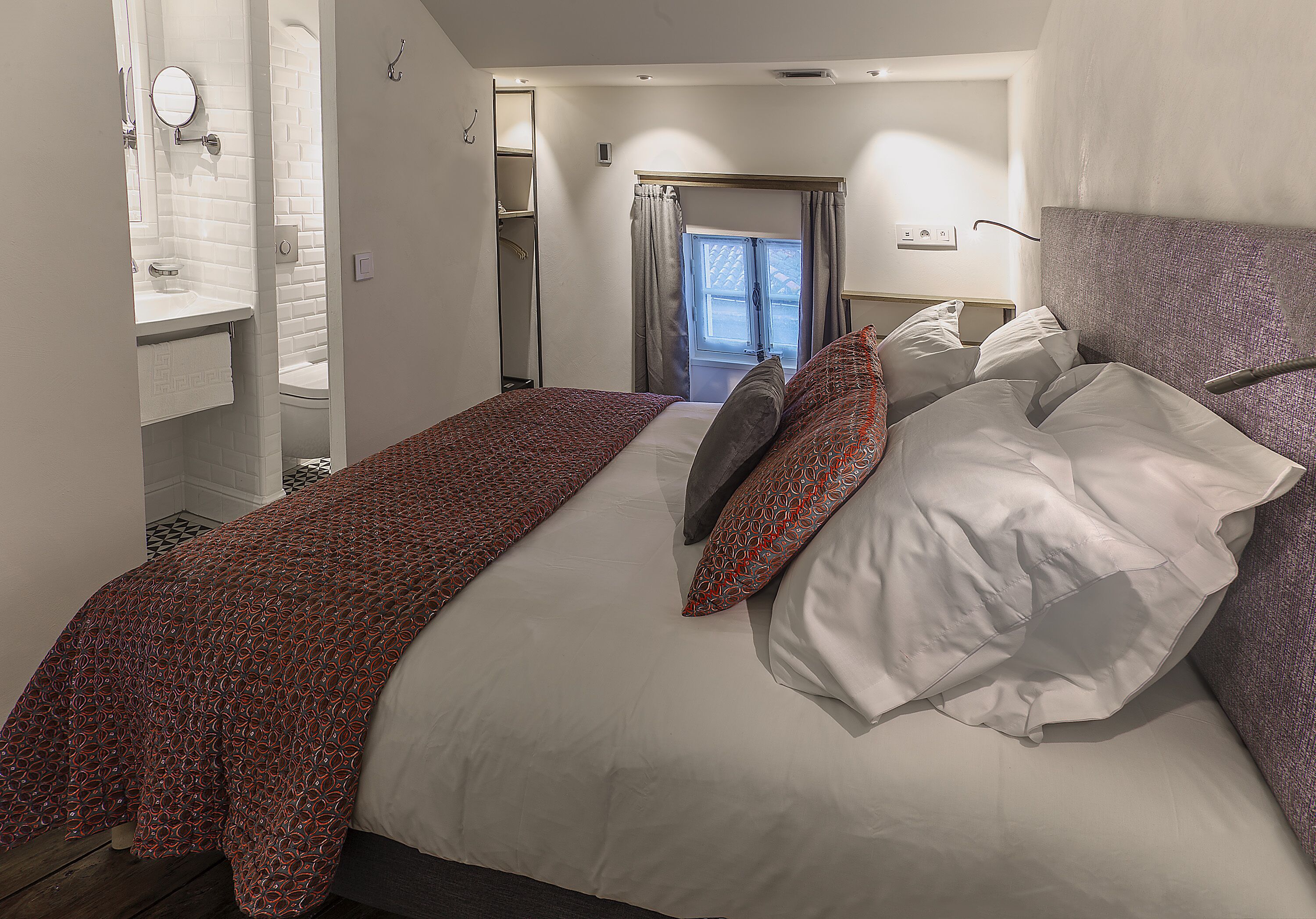 A bedroom with a bed, pillows, a bedside table, a window, and a bathroom visible through an open door.