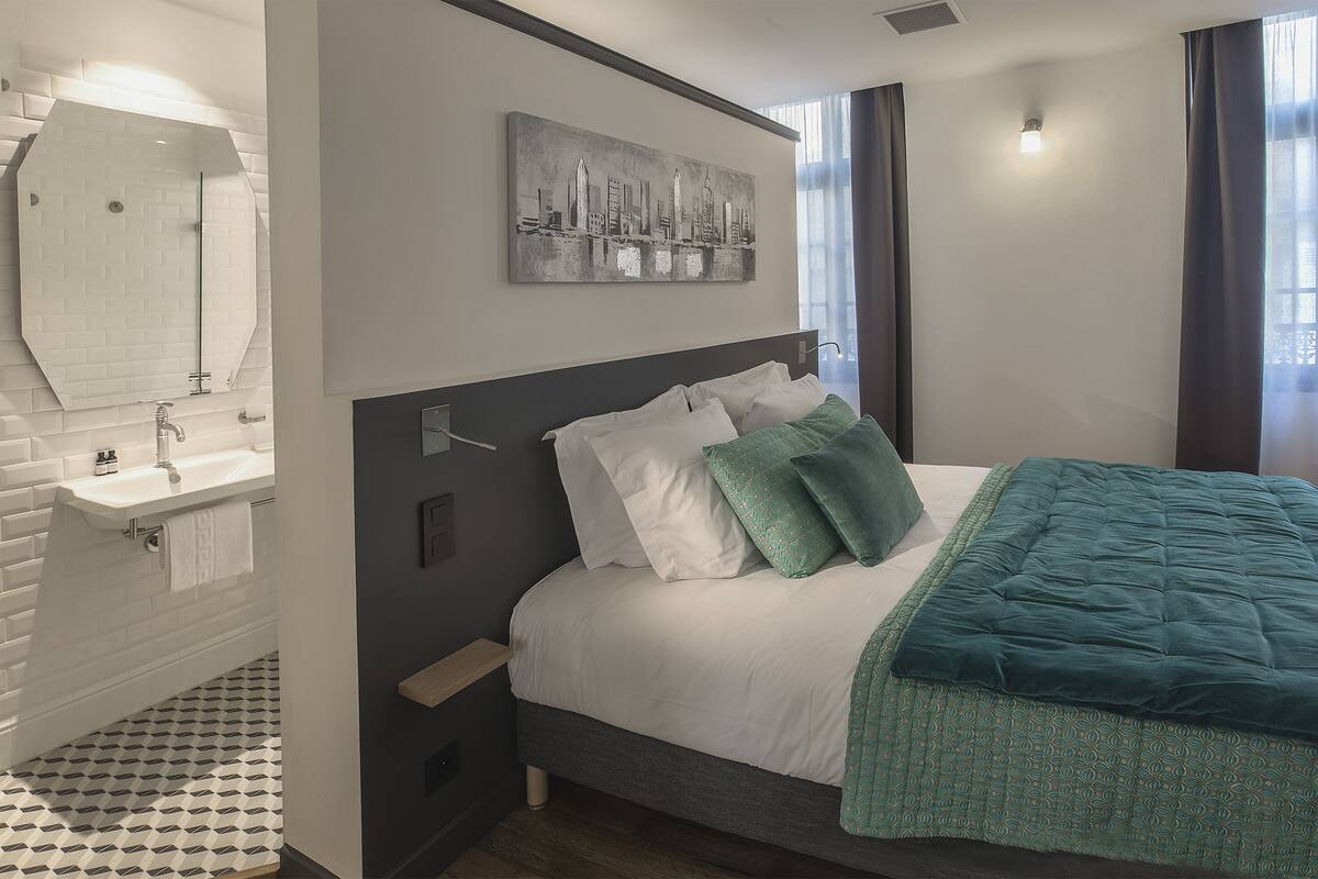 A bedroom with a bed, a nightstand, a wall-mounted mirror, a cityscape artwork, and a bathroom with a sink and tiled floor.