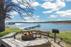 A lakeside deck with a fire pit, wooden benches, and a dock extending into the water.