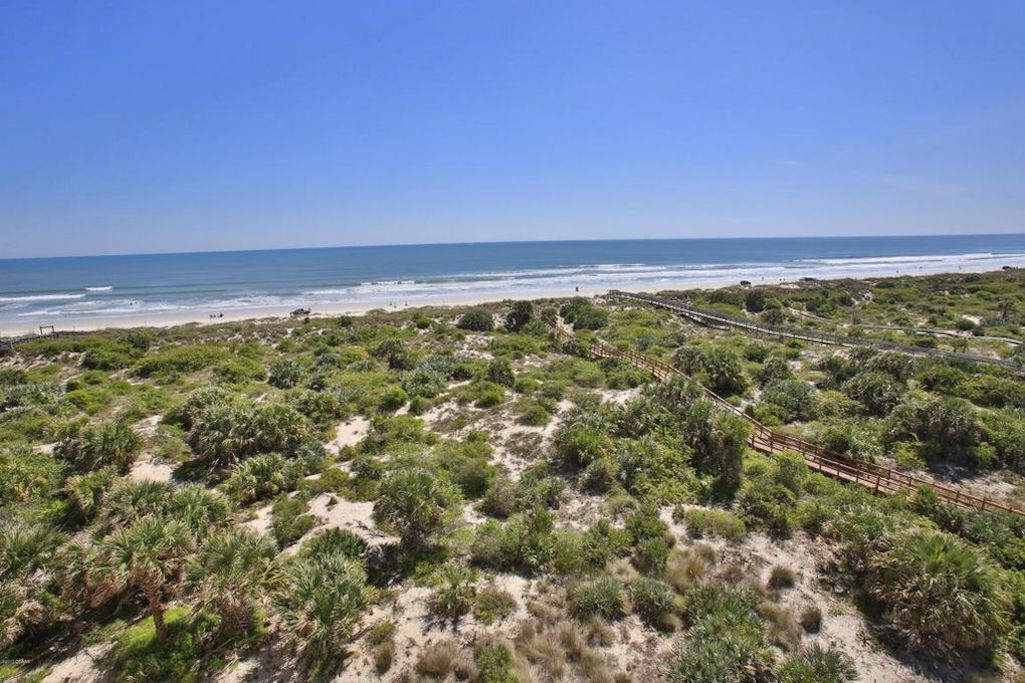 Eine Küstenlandschaft mit Sandstrand, dichter grüner Vegetation und einem hölzernen Steg, der zum Strand führt.