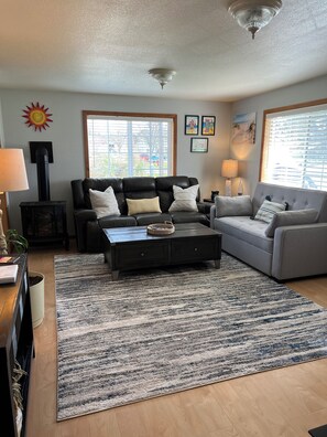 A living room with a black leather sofa, a grey sofa, a coffee table, and a patterned rug. There are two windows with blinds, a ceiling fan, and wall art.
