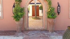 Une porte surmontée d’une arche décorative, flanquée de grands pots de plantes et de lanternes fixées au mur.