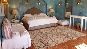 Une chambre avec un grand lit, deux tables de chevet avec des lampes, une petite table sur laquelle est posé un vase, un miroir et un tapis à motifs.