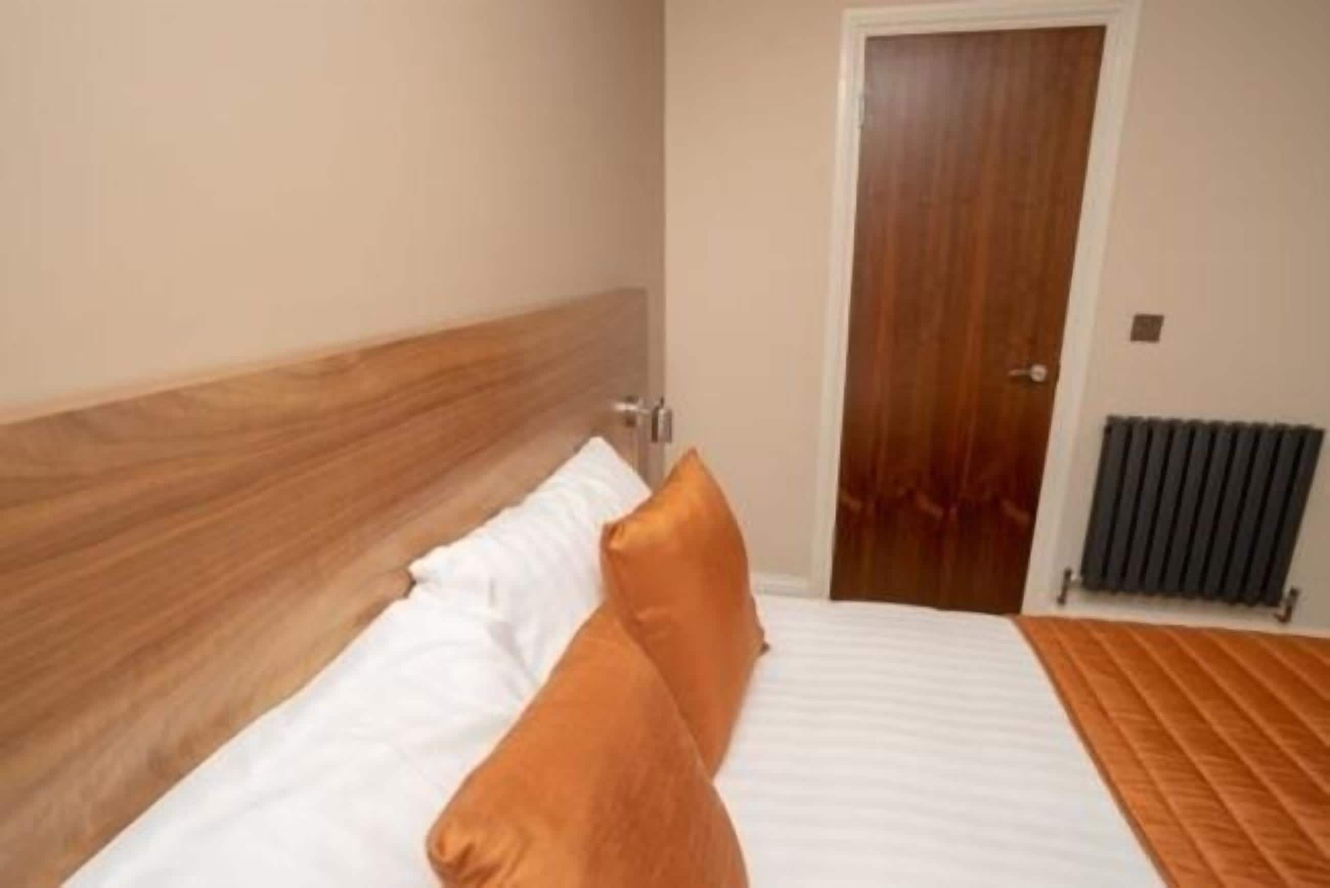 Habitación de hotel con cabecera de madera, cama con ropa de cama blanca y naranja, y una puerta con un radiador al fondo.
