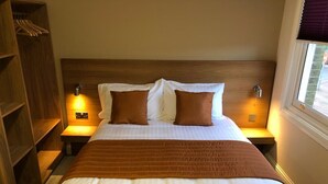 A neatly made bed with a brown blanket, two pillows, and a wooden headboard. There are two bedside tables with lamps, and a wooden wardrobe to the left. - Eagle Hotel (Luton)