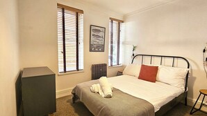 Un dormitorio con cama, un radiador, una ventana con persianas, un cuadro enmarcado en la pared y un taburete pequeño.