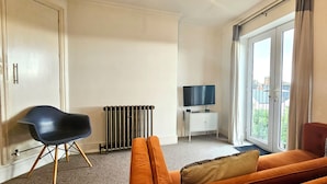 Una habitación con sofá, una silla, un radiador, un televisor y un ventanal corredera al exterior.