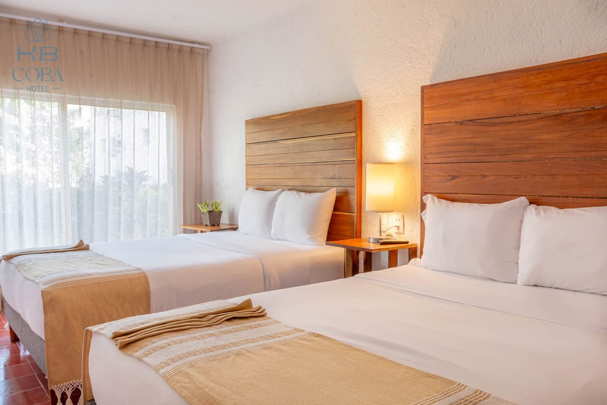 Habitación de hotel con dos camas, cabeceros de madera, una mesita de noche con lámpara y una ventana con cortinas traslúcidas.