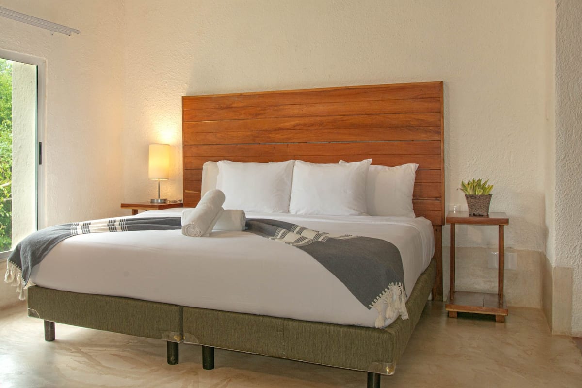 Una cama bien hecha con sábanas blancas y una manta gris, cabecera de madera, mesita de noche con lámpara y una pequeña planta en maceta.