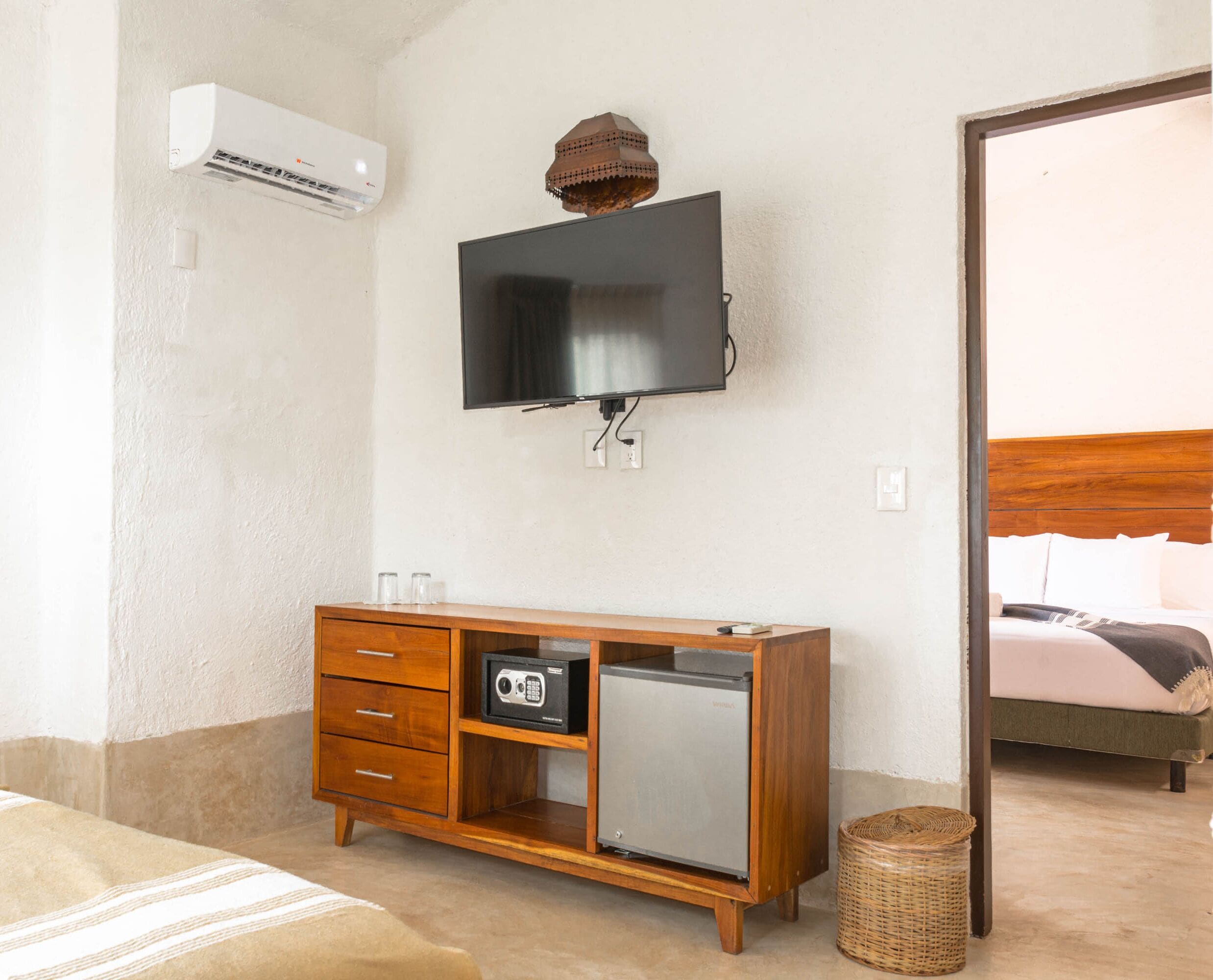 Quarto de hotel com um armário de madeira, televisão fixada na parede, um pequeno frigorífico e uma cesta de vime no chão.