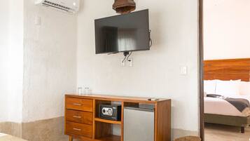 Quarto de hotel com um armário de madeira, televisão fixada na parede, um pequeno frigorífico e uma cesta de vime no chão.