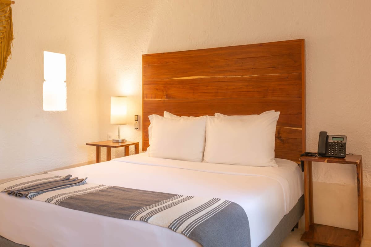 Una habitación de hotel con cabecera de madera, cama con ropa blanca, lámpara de noche, teléfono y una mesita con lámpara.
