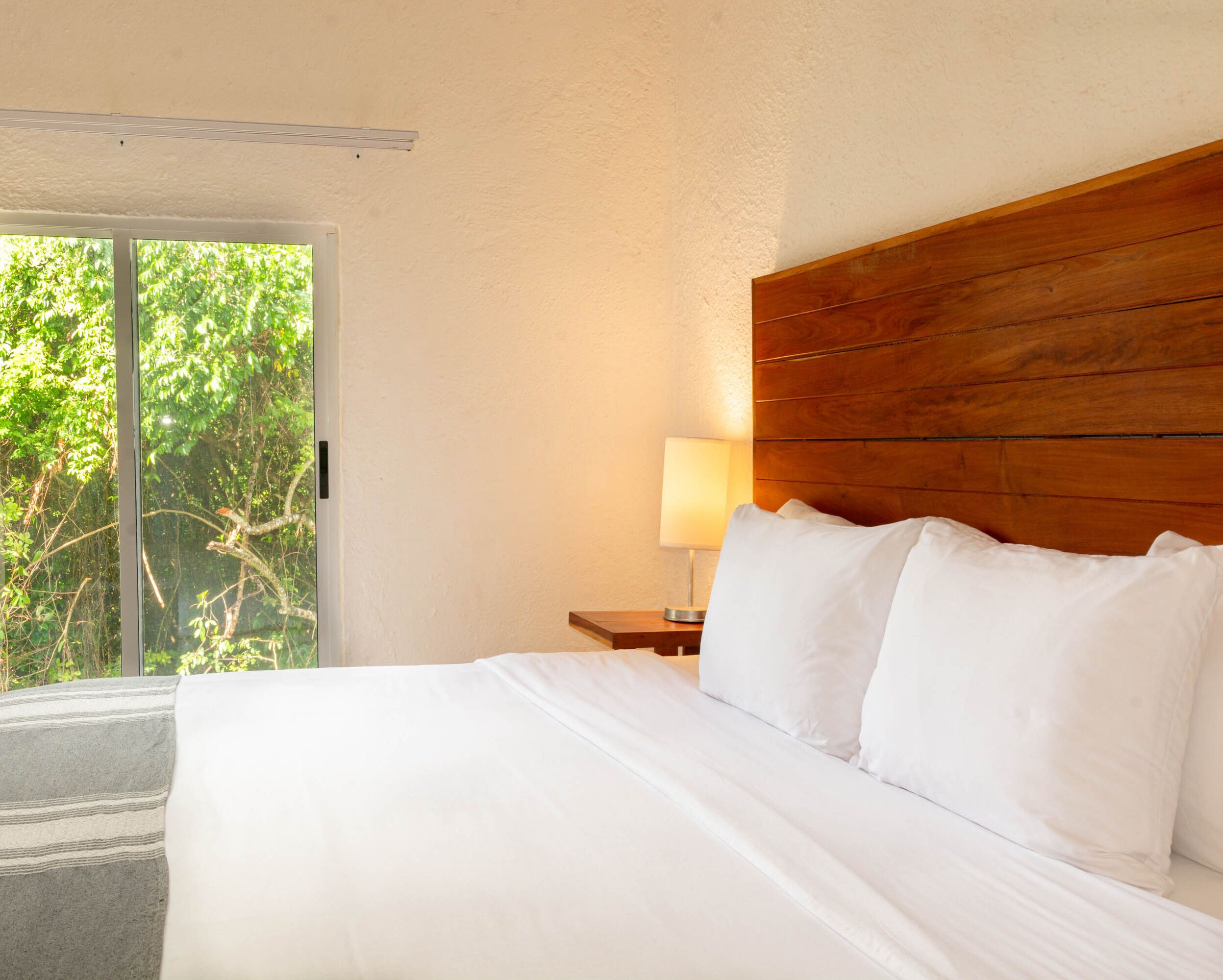 Camera d'albergo con un letto, una testiera in legno, un comodino con una lampada e vista sul verde attraverso una porta scorrevole.