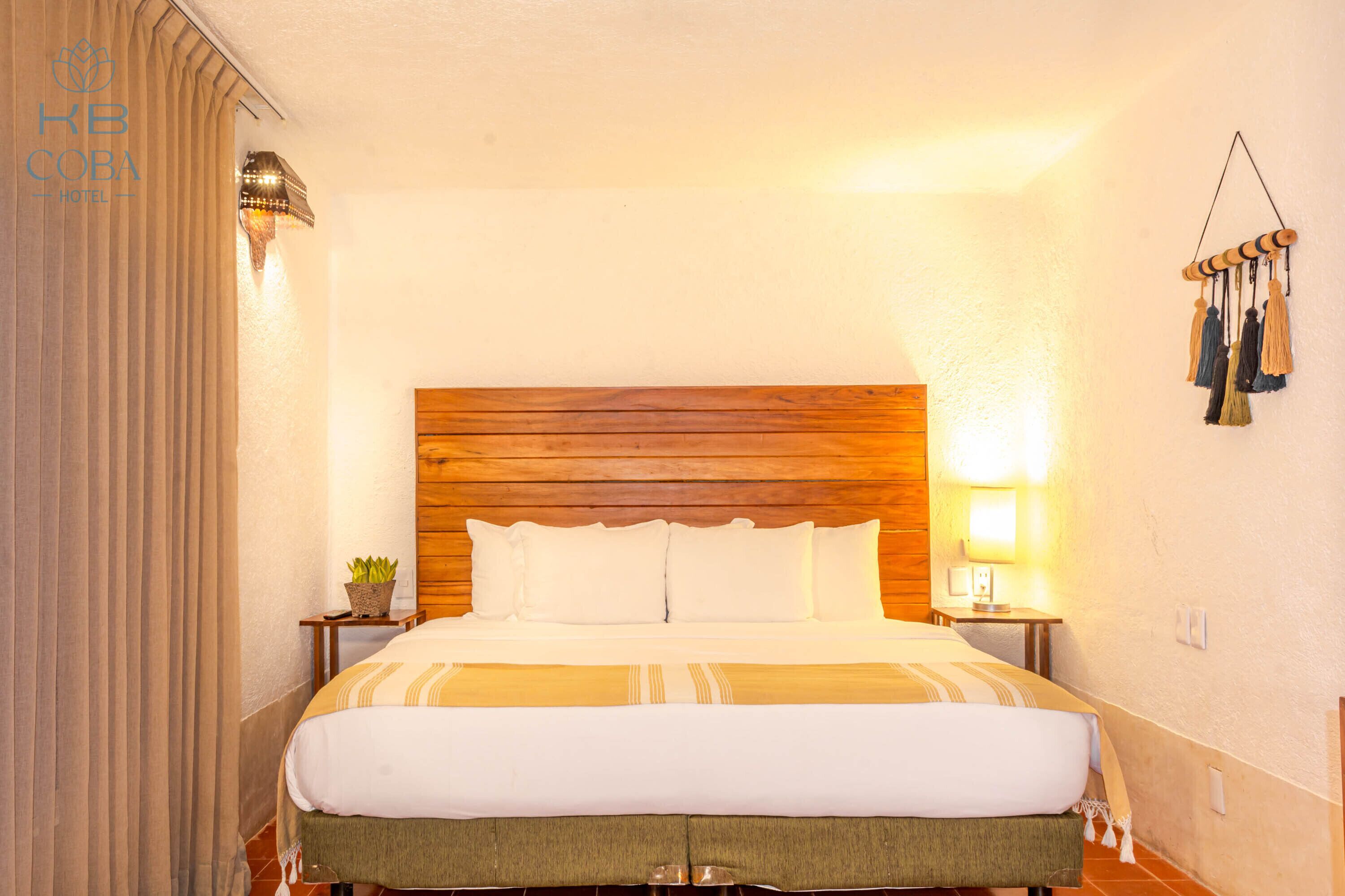 A hotel room with a wooden headboard, a bed with white bedding, a small bedside table with a lamp, and a wall-mounted hanging decoration.