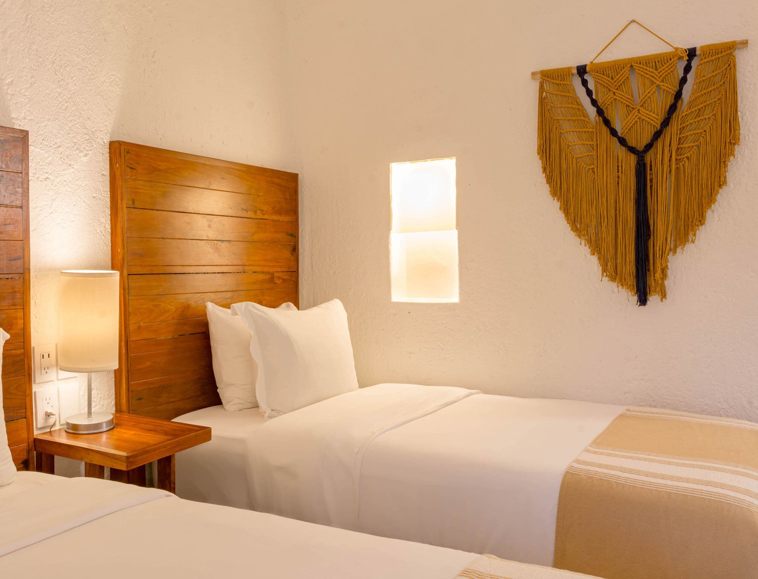 Camera d'albergo con testiera in legno, abat-jour, decorazione macramè a parete e finestra con lampada.