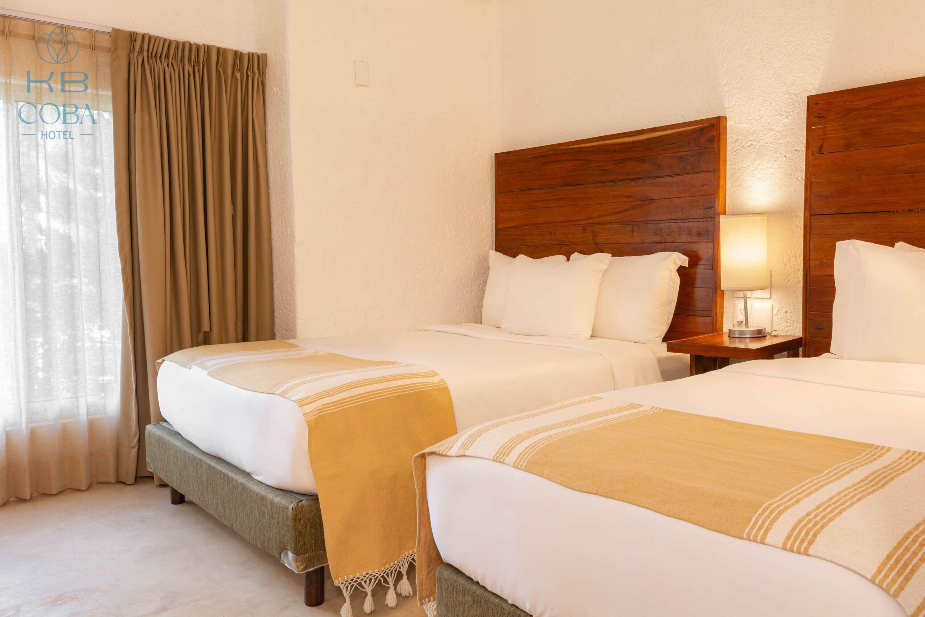 Une chambre d’hôtel avec deux lits, une tête de lit en bois, une table de chevet avec une lampe et une fenêtre avec des rideaux.