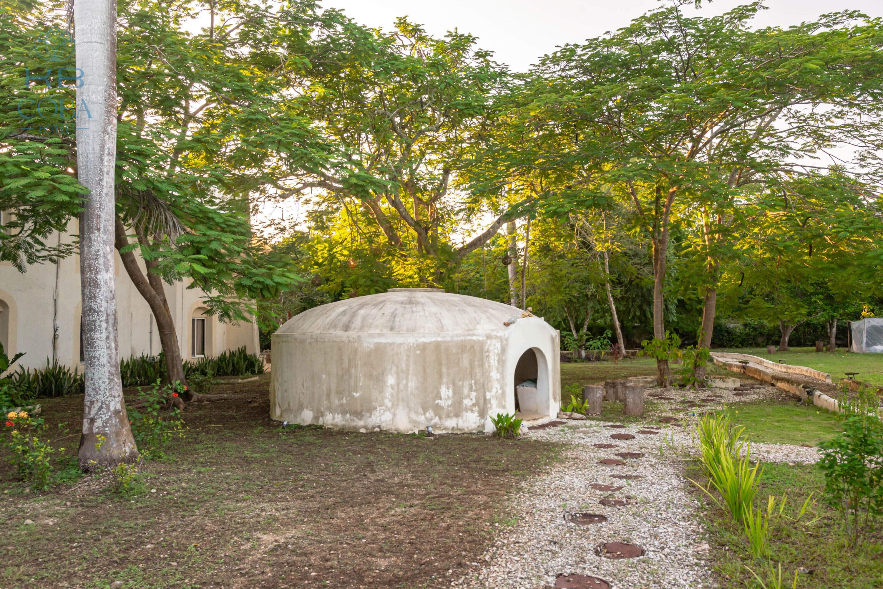 A traditional yurt in a garden setting with a gravel path leading to it, surrounded by trees and plants.