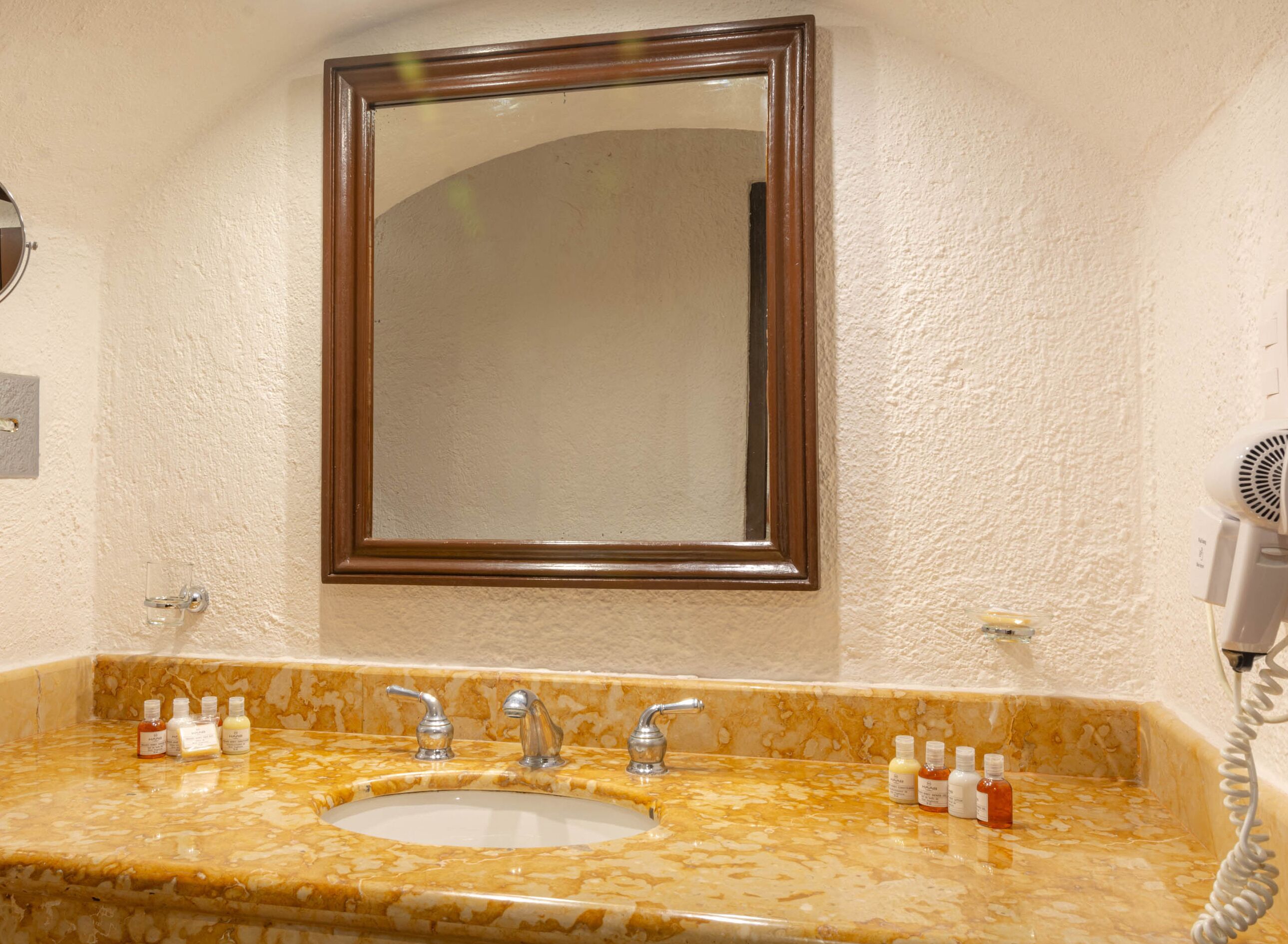 Une salle de bain avec un lavabo en marbre, deux robinets, un miroir et un sèche-cheveux fixé au mur.