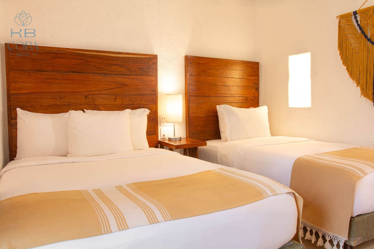 Habitación de hotel con dos camas, cabeceros de madera, ropa de cama blanca, una lámpara de noche y una ventana con vistas.