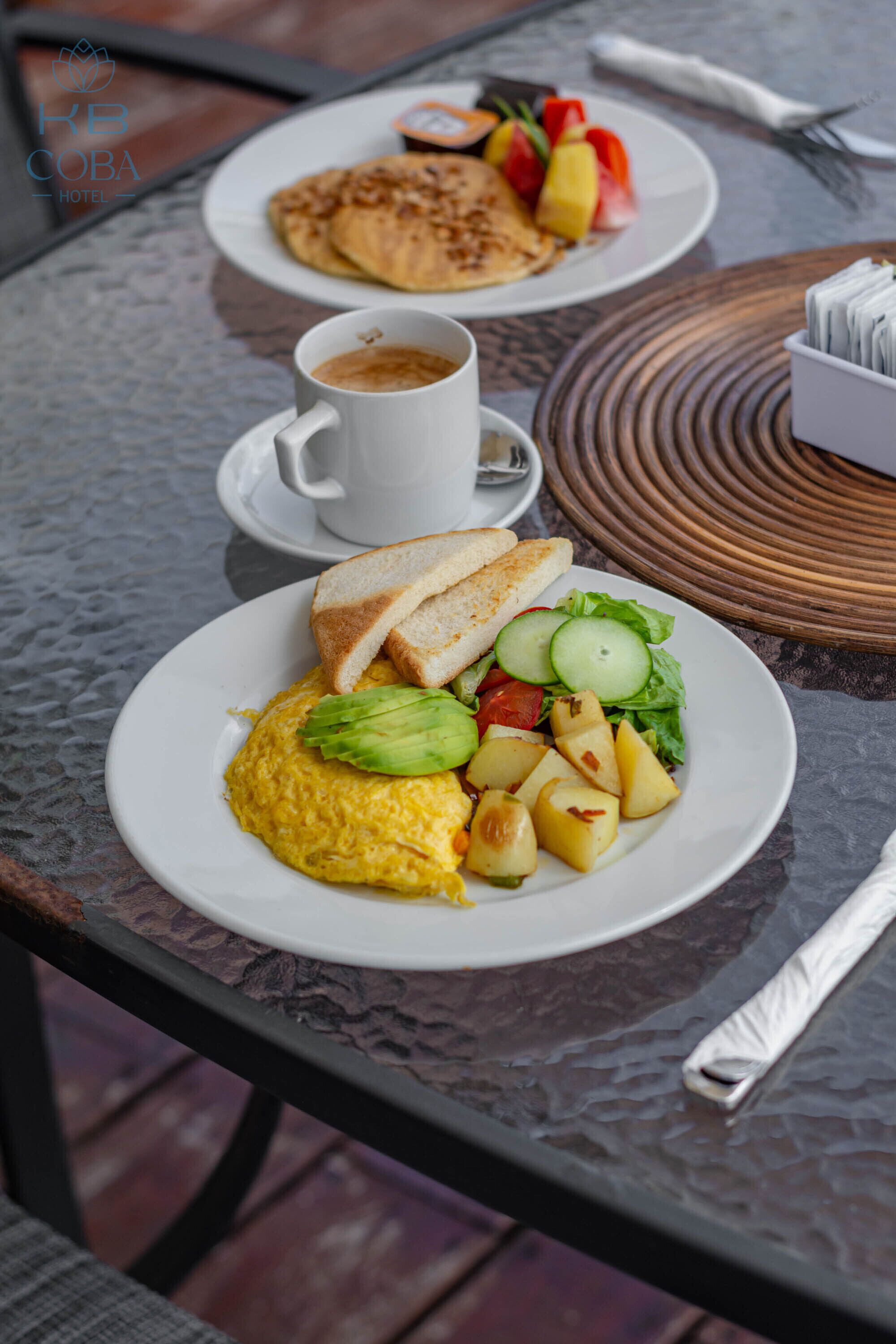 Un desayuno con huevos revueltos, tostadas y verduras, una taza de café y otro plato con tortitas y fruta.