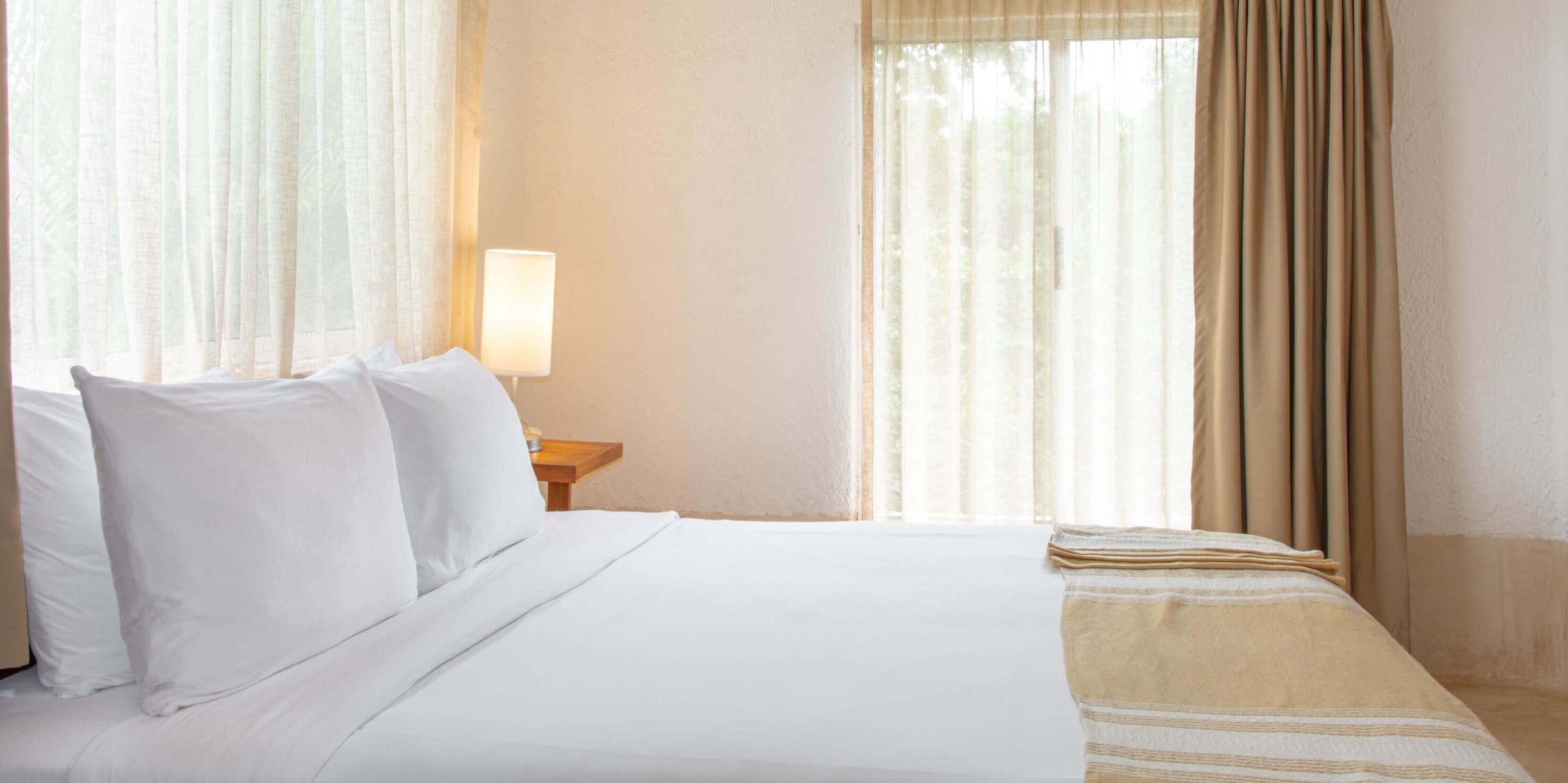A hotel room with a bed, a nightstand with a lamp, and sheer curtains.