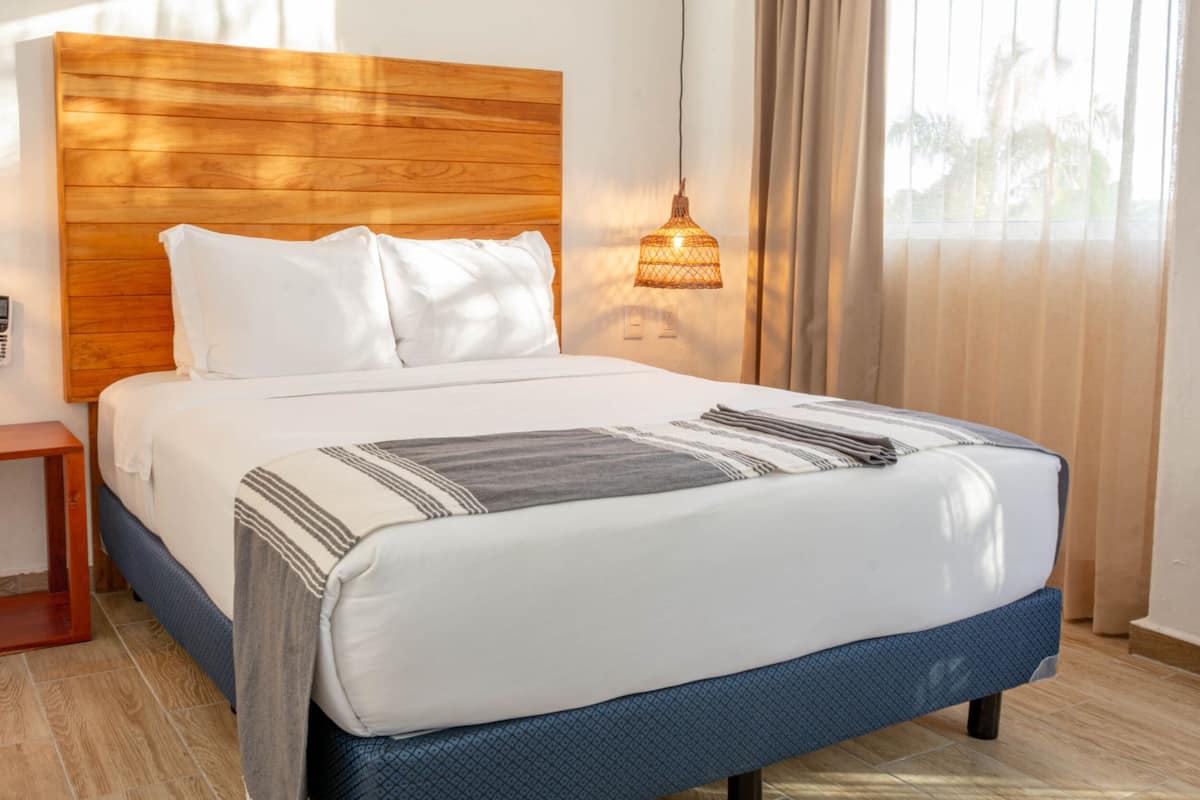 Habitación de hotel con cabecera de madera, cama con ropa blanca y un plaid estampado, mesita de noche y lámpara colgante.
