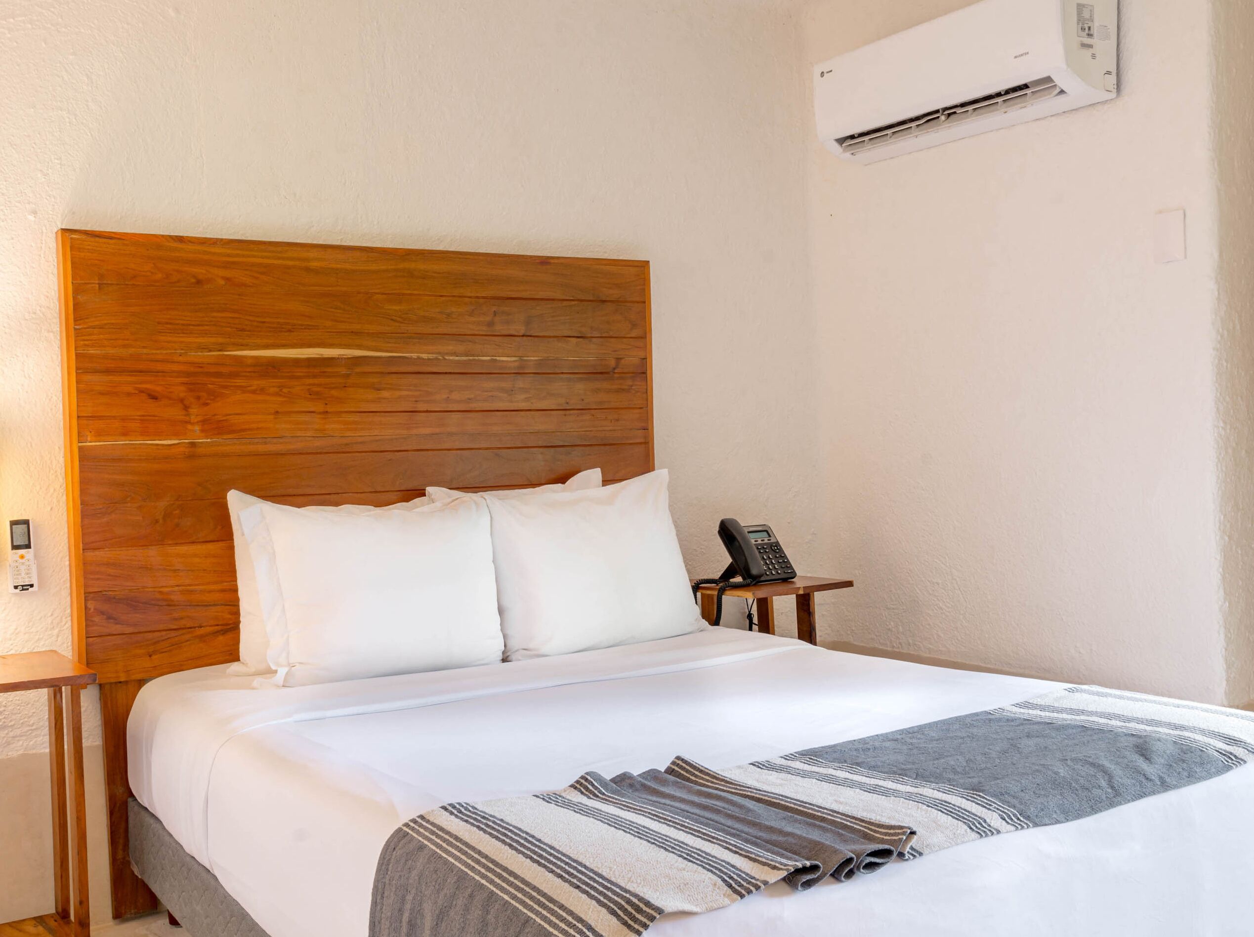 A hotel room with a wooden headboard, a bed with white bedding, a bedside phone, and an air conditioning unit on the wall.