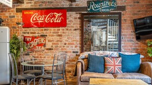 Una habitació acollidora amb una paret de maó, un sofà amb coixins, una taula de vidre amb cadires i un cartell de Coca-Cola.