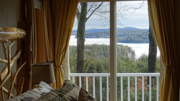 Habitación con vista a un lago y un bosque, cabecera de madera y sofá con cojines estampados.