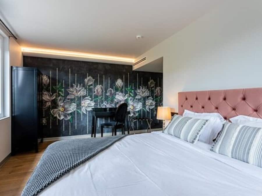 A modern hotel room with a large bed, a desk, and a dark floral wallpaper.