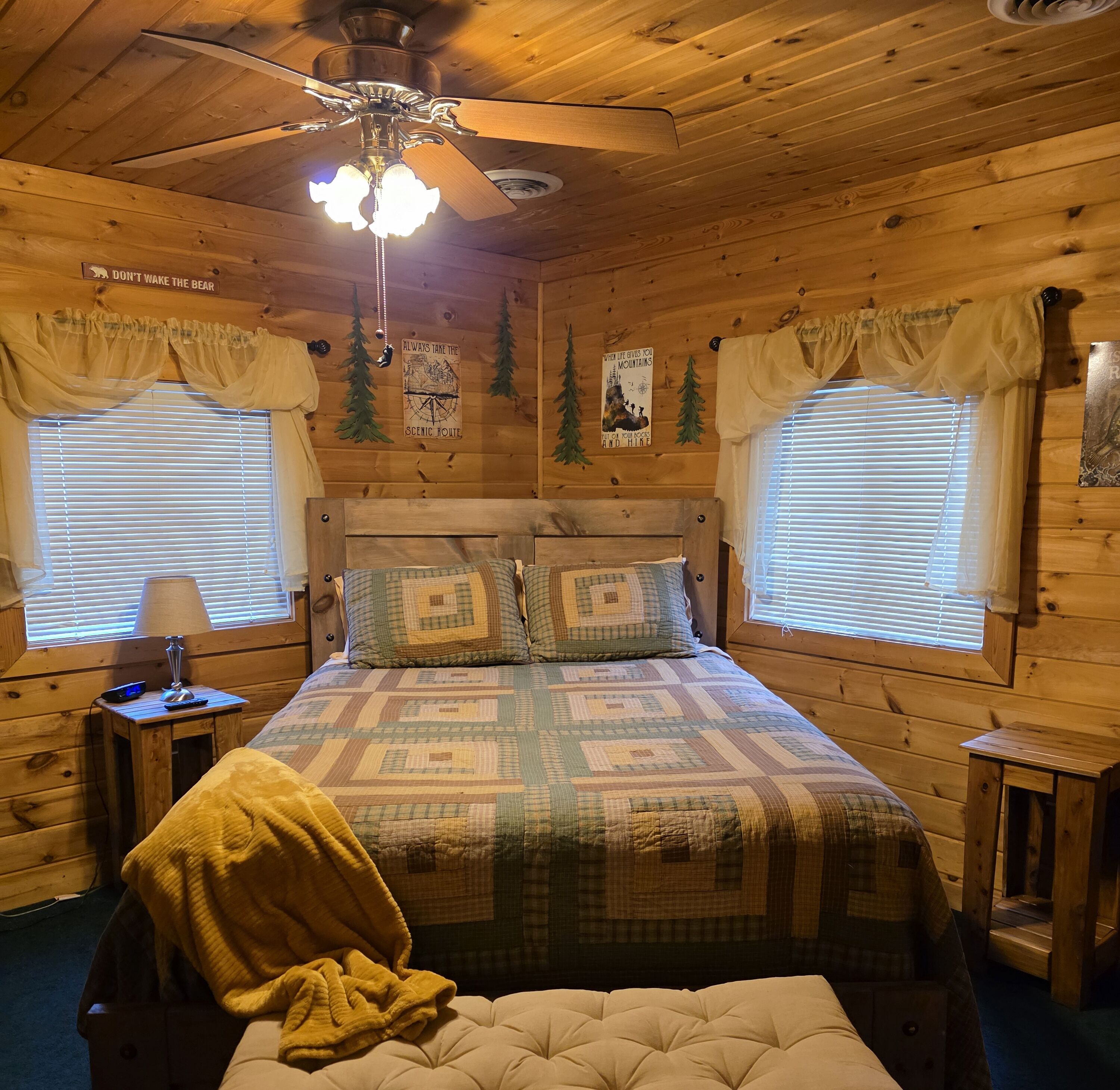 A cozy cabin room with a wooden ceiling, a bed with a quilt, two windows with curtains, a nightstand with a lamp, and a ceiling fan.