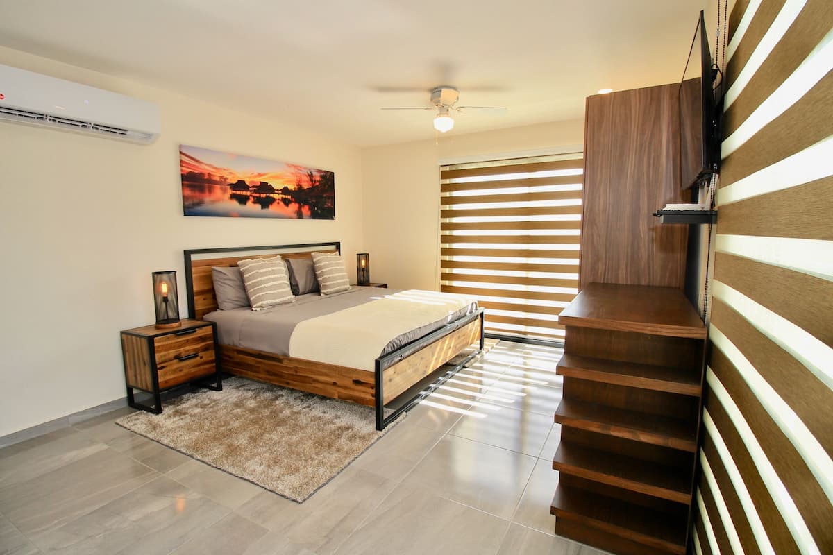 Ein Schlafzimmer mit Bett, Nachttischen, Deckenventilator, Fenster mit Jalousien und einer Holztreppe.