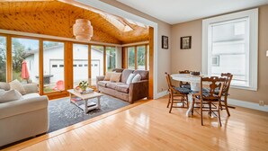 A spacious living room with a wooden ceiling, large windows, a dining area with a table and chairs, and a cozy seating area with a sofa and a coffee table. - The sunroom on Maple  (Picton)