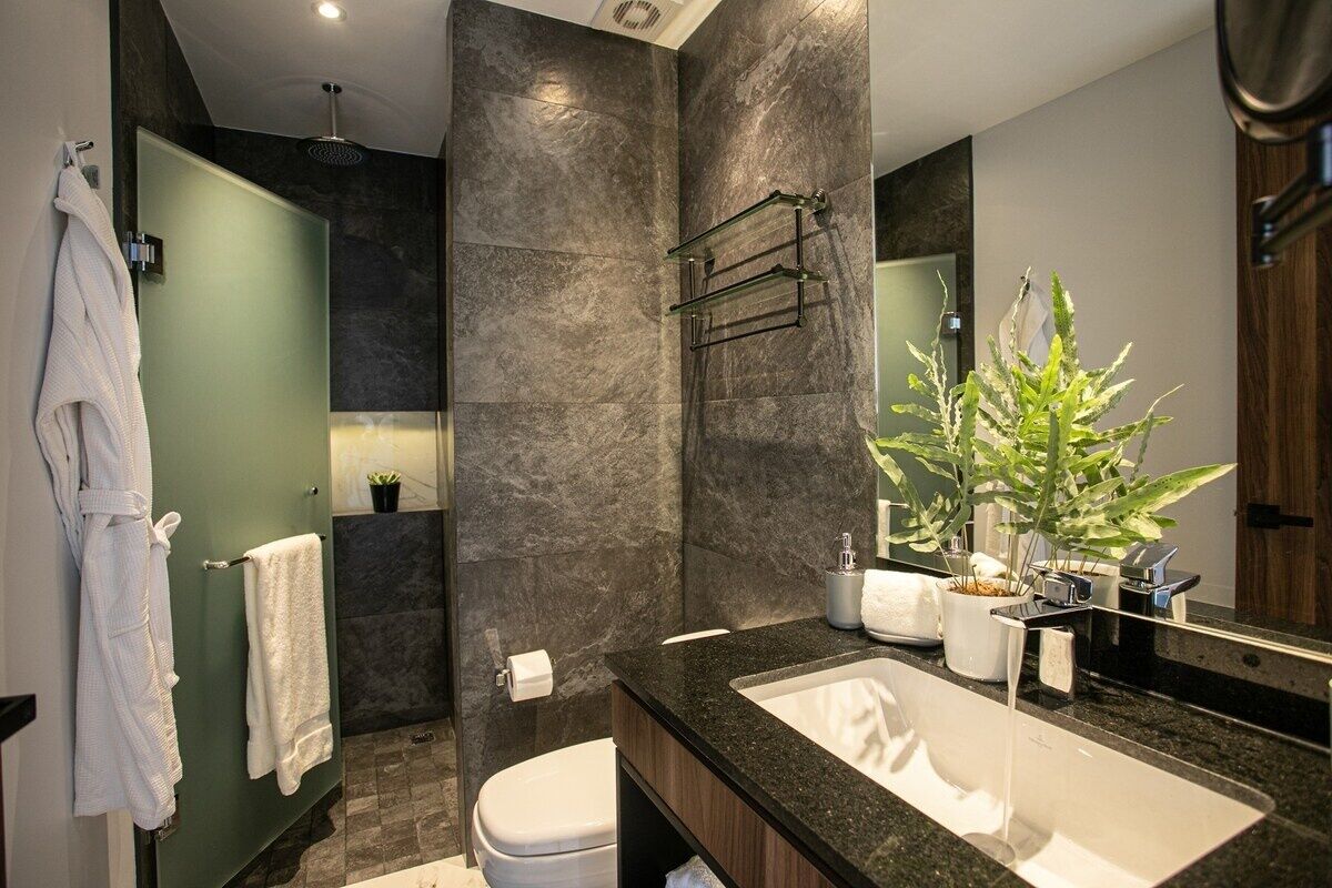 A modern bathroom with a large mirror, dark stone walls, a white sink, and a green door.