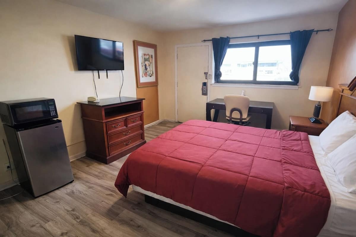 Ein Hotelzimmer mit Bett, Schreibtisch, Stuhl, Fernseher, Kühlschrank und einem Fenster mit Vorhängen.
