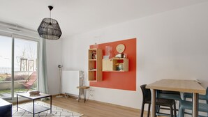 A modern living room with a wooden floor, a dining table with chairs, a bookshelf, a hanging light fixture, and a view through a glass door. - Appartement Corselle - Welkeys (Mouvaux)