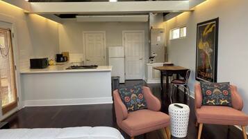 Une chambre moderne avec un lit, deux fauteuils, une petite table et un coin cuisine à l’arrière-plan.