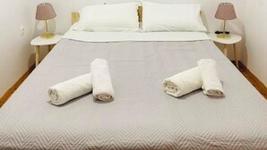 Una cama bien hecha con una colcha blanca y dos toallas enrolladas encima. Hay dos mesitas de noche con lámparas a cada lado de la cama.