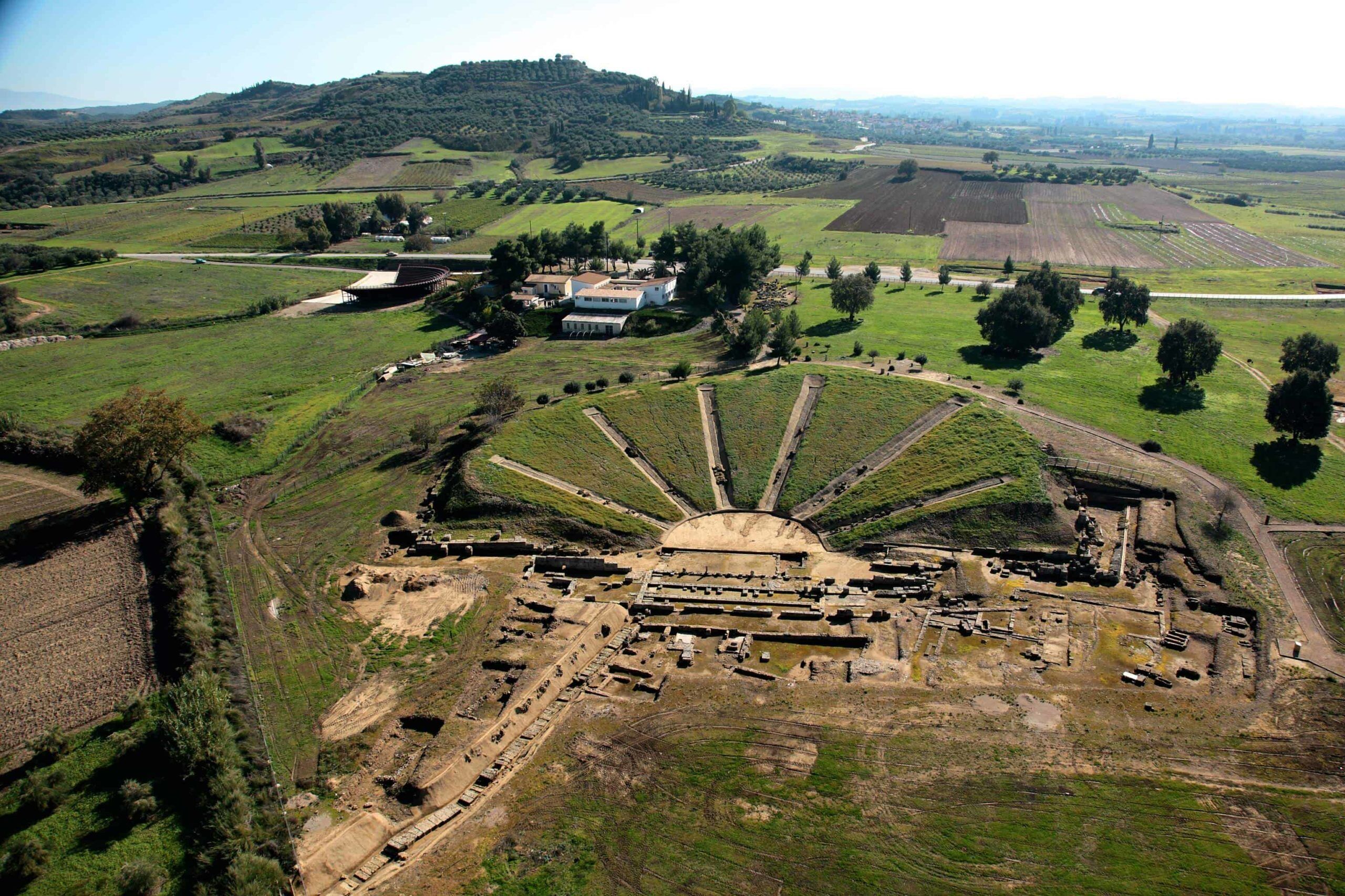 Luchtbeeld van een archeologische site met een centrale, ronde constructie, omringd door terrassen en enkele gebouwen in de verte.