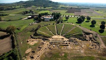Luchtbeeld van een archeologische site met een centrale, ronde constructie, omringd door terrassen en enkele gebouwen in de verte.